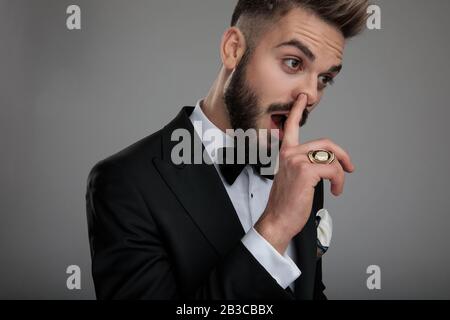 Nahaufnahme eines tollpatschigen Bräutigam, der seine Nase pflückt und seinen Ring zeigt, während er Tuxedo und Bowtie trägt und auf grauem Studiohintergrund steht Stockfoto