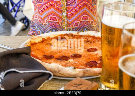 Eine Frau sitzt an einem Café-Tisch im Freien mit Pfefferoni-Pizza und Bier auf dem Tisch vor ihr. Stockfoto