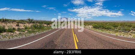Panoramablick auf eine lange, gerade Straße in der Wüste, die zu einer schönen Bergkette unter einem blauen Himmel mit Wolken führt Stockfoto