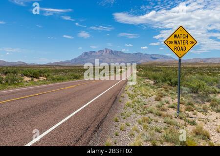 Schild "auf der Straße nach Wasser Ausschau halten" entlang einer langen geraden Straße in der Wüste, die zu einer schönen Bergkette unter einem blauen Himmel mit Wolken führt Stockfoto