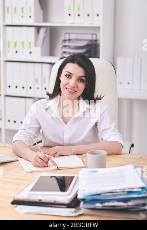 Porträt der lächelnden, attraktiven jungen Frau in weißem Hemd, das am Schreibtisch sitzt und im Büro arbeitet Stockfoto
