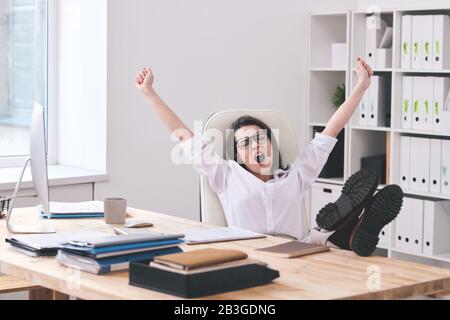Müde junge Bürofrau, die mit Füßen auf dem Tisch sitzt und mit ausgestreckten Armen im Büro gähnelt Stockfoto
