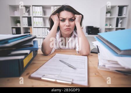 Portrait von frowning junge Geschäftsfrau verwirrt mit Schreibarbeit, die mit Kopf in den Händen am Bürotisch sitzt Stockfoto