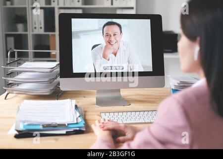 Über die Schulter schauen, wie Geschäftsfrau am Schreibtisch sitzt und Computer verwendet, während sie online mit asiatischen Geschäftspartnern spricht Stockfoto