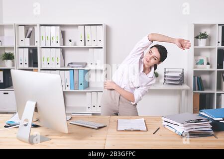 Die energiegeladene junge Frau in der Bluse beugt sich an der Seite und streckt den Arm, während sie im Büro warm-up-Übungen macht Stockfoto