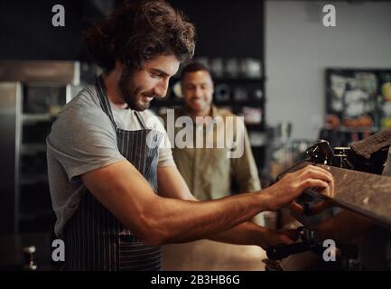 Gut aussehende, erfolgreiche männliche Angestellte, die frischen Kaffee mit der Maschine zubereiten, während der Kunde durch die Theke schaut Stockfoto