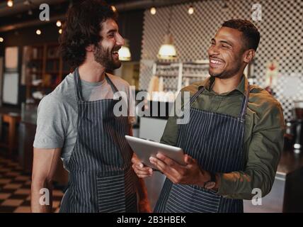 Fröhliche männliche Kollegen, die ein digitales Tablet am Café-Schalter verwenden und lachen Stockfoto