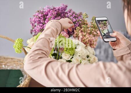 Eine Frau macht ein Foto auf ihrem Telefon mit Blumen. Blumengeschäft Konzept . Blumenfrau schafft Blumenarrangement in einem Korbkorb. . Blumen Lieferung. Stockfoto