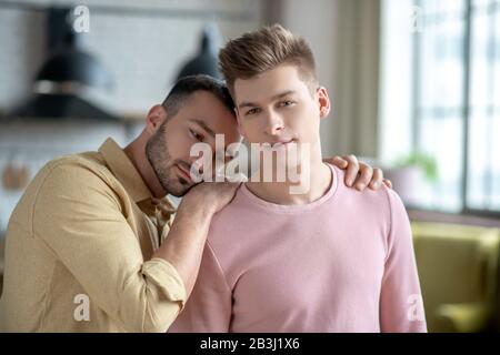 Dunkelhaariger Mann in orangefarbenem Hemd, der die Hände auf die Schulter seiner Partner legt und friedlich aussieht Stockfoto