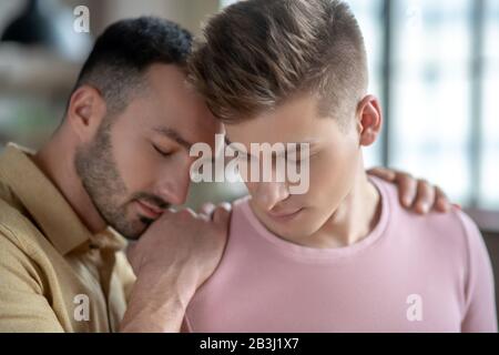 Dunkelhaariger Mann in orangefarbenem Hemd, der die Hände auf die Schulter seiner Partner legt, wobei die Augen geschlossen sind und sich sicher fühlen Stockfoto