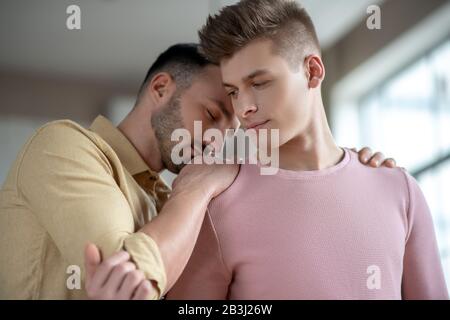 Dunkelhaariger Mann in orangefarbenem Hemd, der die Hände auf die Schulter seiner Partner legt, wobei die Augen geschlossen sind Stockfoto