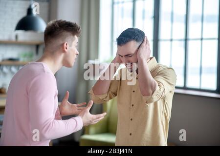 Mann in orangefarbenem Hemd, der seine Ohren schließt, während sein Partner ihn anschreit Stockfoto