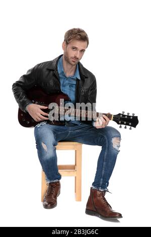 Charmanter Casual man mit schwarzer Lederjacke sitzt auf einem Holzstuhl, spielt leidenschaftlich Gitarre und blickt auf weißen Studiohintergrund Stockfoto