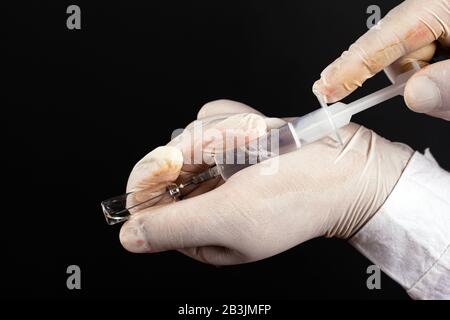 Der Arzt sammelt Medikamente aus einer Ampulle in einer Spritze mit weißen sterilen Handschuhen auf dunklem Hintergrund in Nahaufnahme. Impfspritzmittel Agai Stockfoto