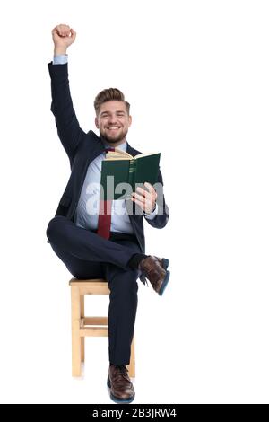 Begeisterter junger Geschäftsmann lächelt, hält Hand in die Luft und liest ein Buch, sitzt isoliert auf weißem Hintergrund im Studio Stockfoto