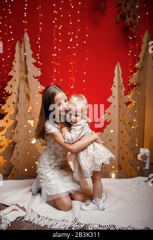 Zwei kleine Schwestern umarmen sich vor dem Hintergrund roter Weihnachtsdekorationen, Idee und Konzept guter und freundlicher Beziehungen in der Familie Stockfoto