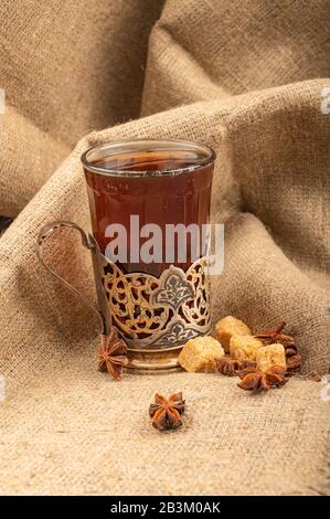 Ein Glas Tee in einem Vintage-Cup-Halter, Sternanis und braunem Zucker auf dem Hintergrund rauer Homespun-Qualität. Nahaufnahme Stockfoto