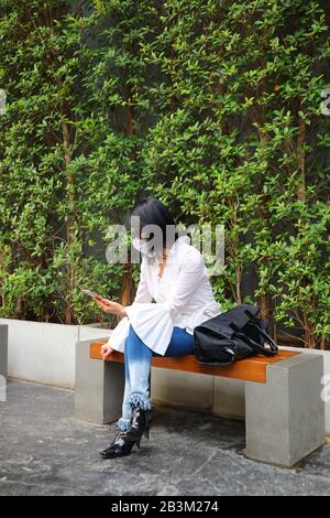 Frau, die Gesichtsmaske trägt, um zu verhindern, dass die Grippe auf einer Bank im öffentlichen Park sitzt Stockfoto