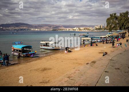 Aqaba, JORDANIEN - 31. JANUAR 2020: Lokale Menschen und Touristen genießen Freitag am sonnigen Stadtstrand mit Glasbodenbooten Urlaub. Winter puffige Wolken Morgenhimmel. Rotseegulf, Haschemitisches Königreich Jordanien Stockfoto