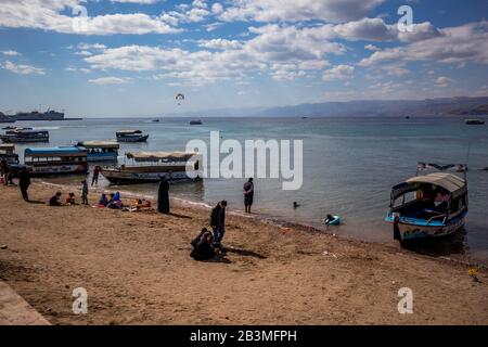 Aqaba, JORDANIEN - 31. JANUAR 2020: Lokale Menschen und Touristen genießen Freitag am sonnigen Stadtstrand mit Glasbodenbooten Urlaub. Winter puffige Wolken Morgenhimmel. Rotseegulf, Haschemitisches Königreich Jordanien Stockfoto