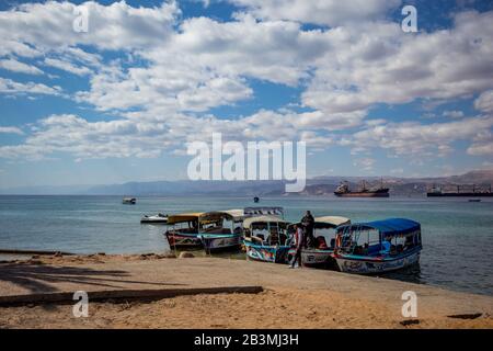 Aqaba, JORDANIEN - 31. JANUAR 2020: Einheimische Männer, Führer mit Glasbodenbooten warten neugierige Touristen, Urlaub Freitag am sonnigen Stadtstrand. Winter puffige Wolken Himmel. Rotseegulf, Königreich Jordanien Stockfoto