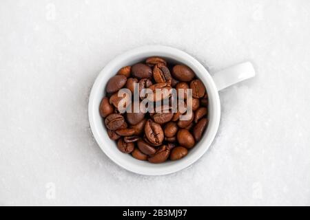 Eine weiße Tasse mit frisch gerösteten Kaffeebohnen auf Schneegrund. Nahaufnahme, selektiver Fokus, Draufsicht Stockfoto