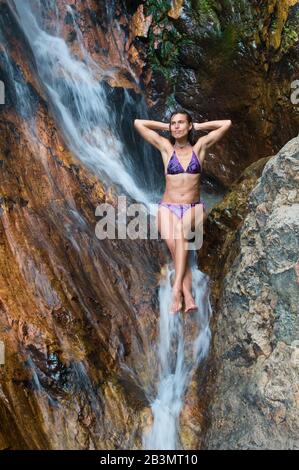 Die Frau macht sich Freude daran, im Wasserfall baden zu können. Junge Frau ist mit violettem zweiteiligem Badeanzug bekleidet. Stockfoto