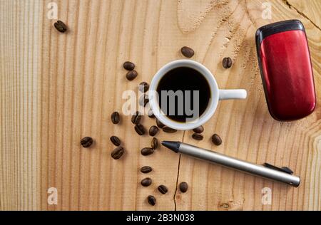 Tasse mit schwarzem Kaffee, Kugelschreiber und altmodischem Handy auf Holzbrett mit wunderschönem Getreide und dargestellten Kaffeebohnen Stockfoto