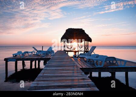 Die aufgehende Sonne umrahmt von einer mit Palmblatt strohgedeckten Cabana und Silhouette eines Stuhls und zwei Hängematten am Ende eines Holzsteges an einem Strand auf Caye Calker, Stockfoto