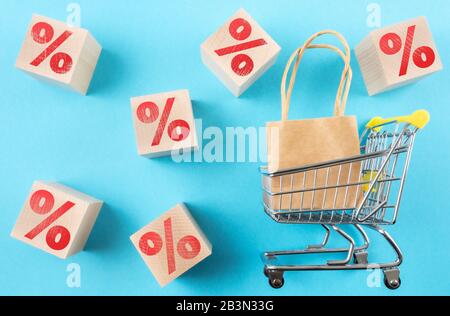Einkaufswagen mit Einkaufstasche und rotem Prozentzeichen auf Holzwürfeln vor blauem Hintergrund, Einzelhandelsverkauf und Rabattkonzept Stockfoto