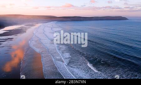 Luftaufnahme des Woolacombe Strandes im Morgengrauen, Wellen, die an einem Strand brechen, an dem die sonnenbeleuchteten Wolken im stehenden Wasser reflektiert werden. Vorgewende am horiz Stockfoto