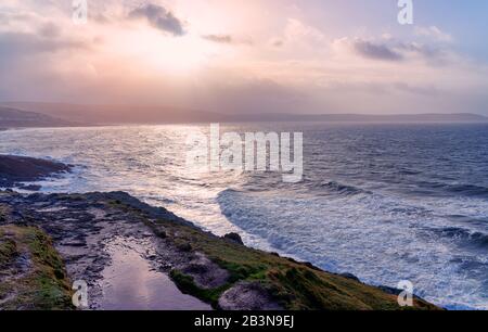 Die Sonne durchbricht Sturmwolken und reflektiert Wellen, die am Ufer brechen, das Küstendorf Woolacombe, das gerade in der Ferne zu sehen ist Stockfoto