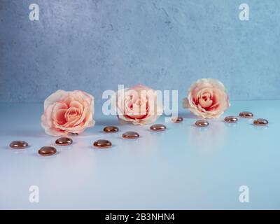 3 pfirsichfarbene Rosen, die mit dunkelorange-braunen, flachen Murmeln aus Glas auf einer weißen Oberfläche mit Pflasterhintergrund aufgereiht sind. Weich und elegant mit dem Refl Stockfoto