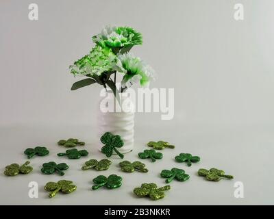 Weiße und grüne Nelken in einer weißen Vase umgeben von glitzernden grünen Vierblättrigen Kleeblättern für St. Patrick's Day Irish Holiday. Stockfoto