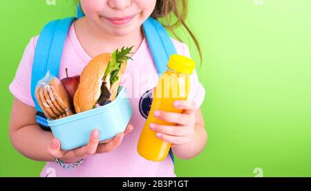 Junge Schulmädchen mit Lunchbox auf grünem Hintergrund. Kleines Mädchen mit Schulrucksack und einem Set Essen für einen Snack. Freier Speicherplatz für Text Stockfoto