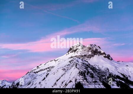 Rosafarbene Wolken liegen in einem blauen Himmel über einem schneebedeckten Alpengipfel. Stockfoto
