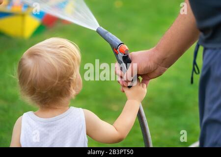 Vater mit Sohn zusammen Bewässerung Garten und Grünrasen zu Hause Hinterhof draußen am heißen Sommertag. Männliche Person mit kleinem Kleinkind Stockfoto