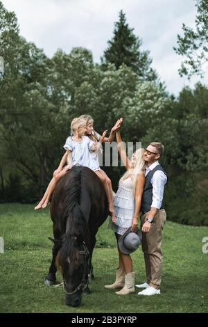 Glückliche Familie auf der Pferderanch, die mit Pferd spazieren geht. Junge glückliche Familie, die sich im Freien auf dem Land lustig macht, zwei Mädchen reiten Pferd, Mutter und Vater Stockfoto