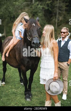 Mutter und Vater gehen mit ihren zwei kleinen niedlichen Töchtern auf dem Pferd in der Natur, auf dem Land. Fröhliche Familie, die auf einem Spaziergang mit braunem Pferd Spaß hat Stockfoto