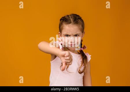 Süßes kleines Mädchen im Kleid mit den Daumen nach unten auf gelbem Hintergrund. Stockfoto