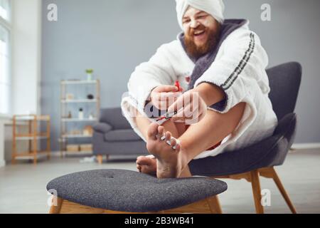 Lustiges Pediküre-Konzept. Lustiger fetter Mann im bademantel und ein Handtuch malt seine Nägel Stockfoto