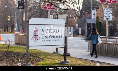 Victoria University (an der Universität von Toronto) Logo auf einem Schild vor dem Campus in der Innenstadt von Toronto, als eine Frau am Bürgersteig geht. Stockfoto