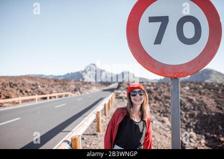 Lifestyle-Porträt einer Frau in Rot auf dem Straßenrand in der Nähe des riesigen Straßenschildes, während Sie auf dem vulkanischen Tal unterwegs sind. Sorgloses Lifestyle- und Reisekonzept Stockfoto