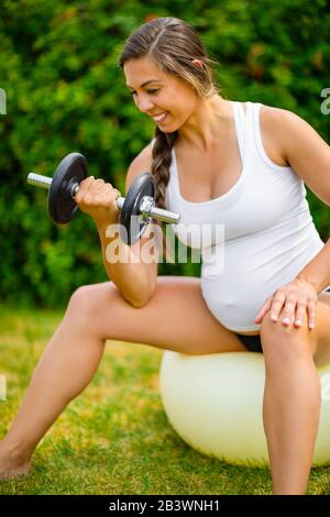 Gesunde Schwangere Frau, Die Hanteln Auf Dem Sportball In Park Hebt Stockfoto