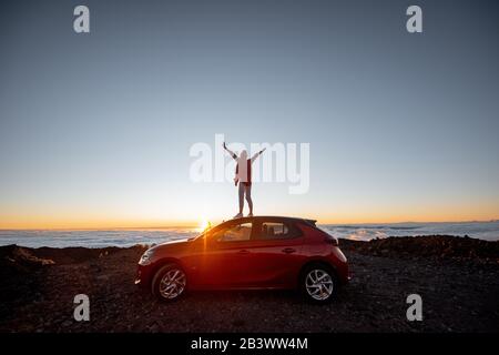 Frau, die bei Sonnenuntergang felsige Landschaften über den Wolken genießt und auf dem Auto hoch in den Bergen steht. Sorgloses Lifestyle- und Reisekonzept Stockfoto