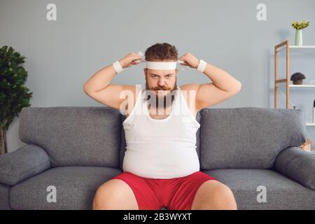 Lustiger fetter Mann in Sportbekleidung sitzt auf dem Sofa im Zimmer Stockfoto