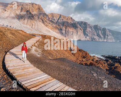Frau, die auf dem malerischen Holzweg durch das felsige Land mit Bergen auf dem Hintergrund spazieren geht. Bild auf Handy gemacht Stockfoto