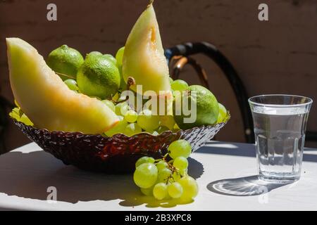 Glas Wasser und Teller mit Obst - Trauben, Zitronen, Melonenscheiben auf dem Tisch an einem sonnigen Tag. Stockfoto