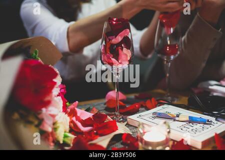 Wunderschönes Glas mit Rosenblättern, Rosenblättern, Rosenblättern an einem romantischen Datum mit Wein, Kerzen im Kerzenlicht und mit feiernden Paaren auf der Rückseite Stockfoto