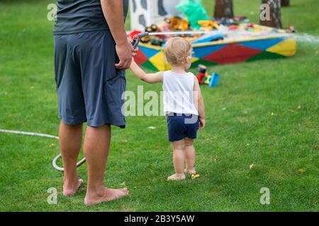 Vater mit Sohn zusammen Bewässerung Garten und Grünrasen zu Hause Hinterhof draußen am heißen Sommertag. Männliche Person mit kleinem Kleinkind Stockfoto